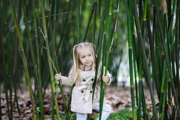 Beautiful little girl with blonde hair standing in a bamboo forest and shaking bamboo