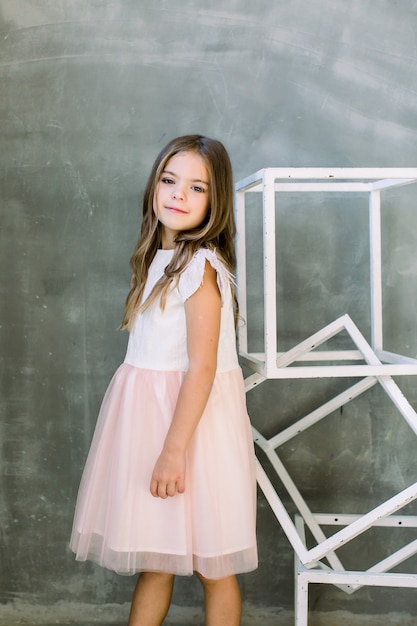 Photo beautiful little girl with brown long hair on a grey wall near the white constructure of the cubes