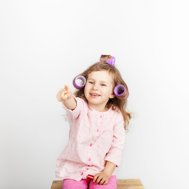 Beautiful little girl with curlers on her head looking at camera and having fun at home