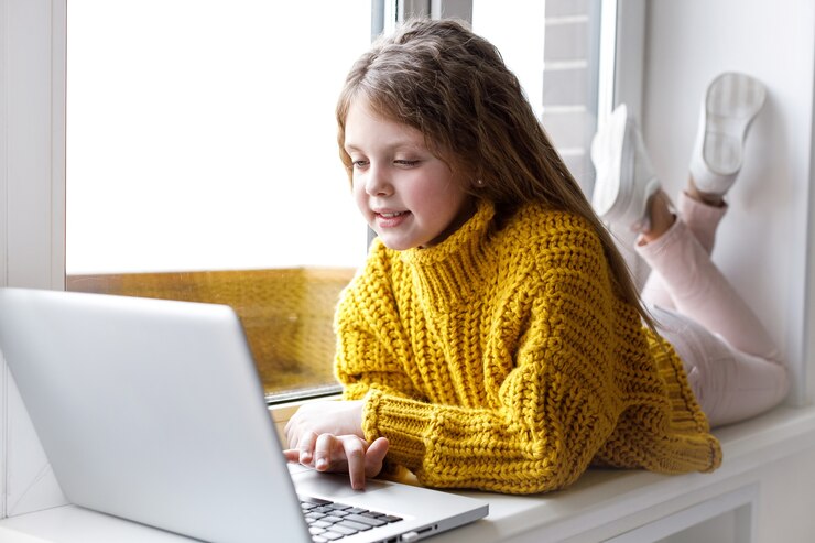 Photo a beautiful little girl with a laptop at home at the window looks at the screen and smiles