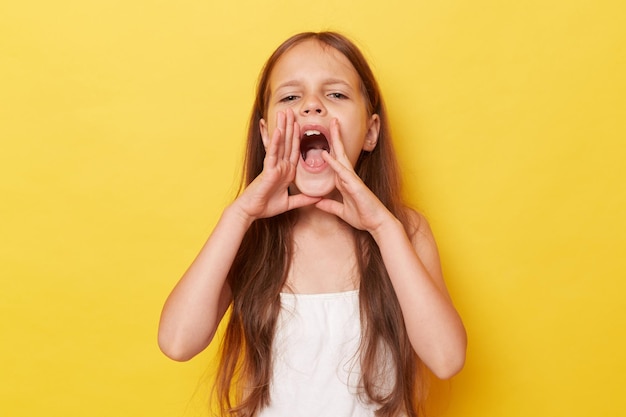 Beautiful little girl with long hair standing isolated over yellow background keeping hands near mouth screaming loud making announcement