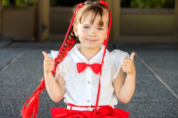 赤いおさげと蝶ネクタイを持つ美しい少女。子供は親指を立てます。高品質の写真