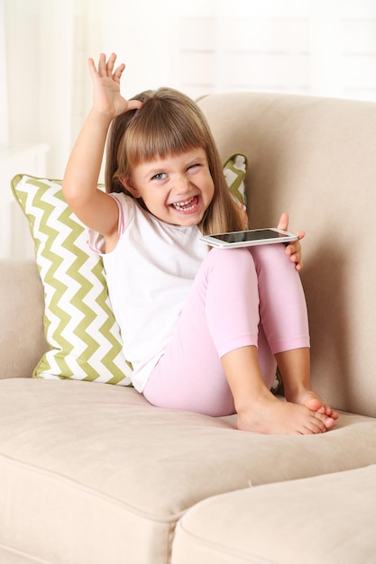 Beautiful little girl with smart phone sitting on sofa on home interior background