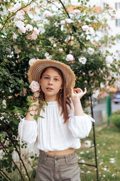 Bella bambina con un cappello di paglia vicino a un cespuglio di rose bianche