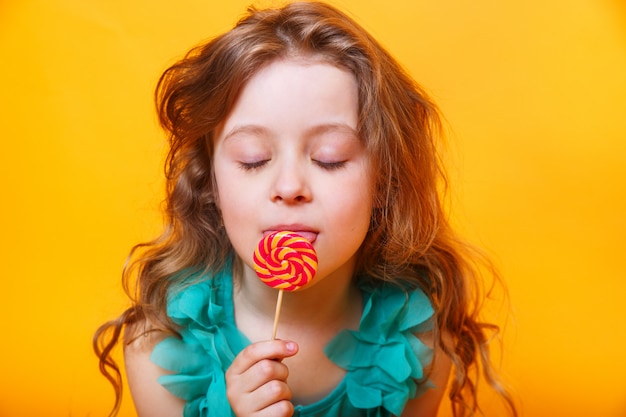 Красивая маленькая девочка с сахарным леденцом на палочке на желтом фоне