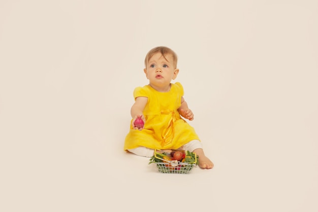 a beautiful little girl in a yellow dress is sitting next to a small basket with vegetables