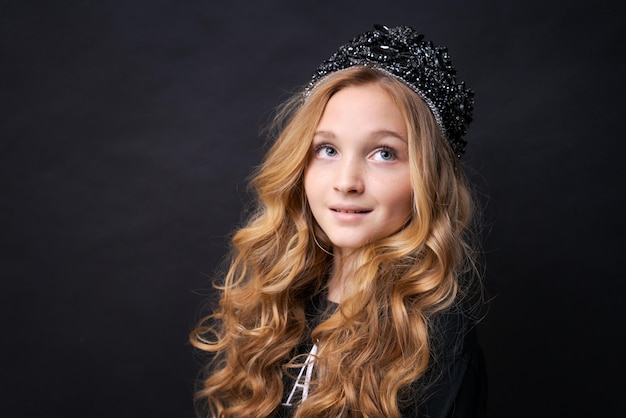 Beautiful little princess girl in black clothes with a crown on her head posing dreamily on a black