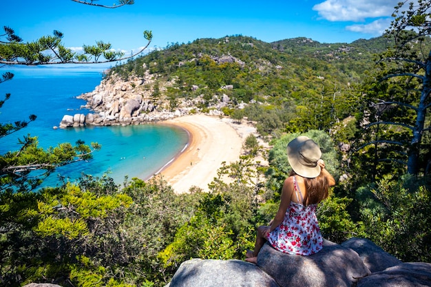 beautiful long-haired girl in dress and hat sits on top of mountain overlooking paradise beach