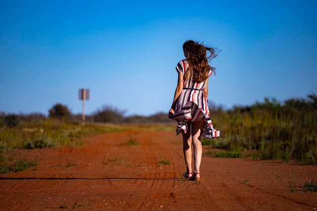 A beautiful long-haired girl in a long dress walks along a road\
in the desert in western australia
