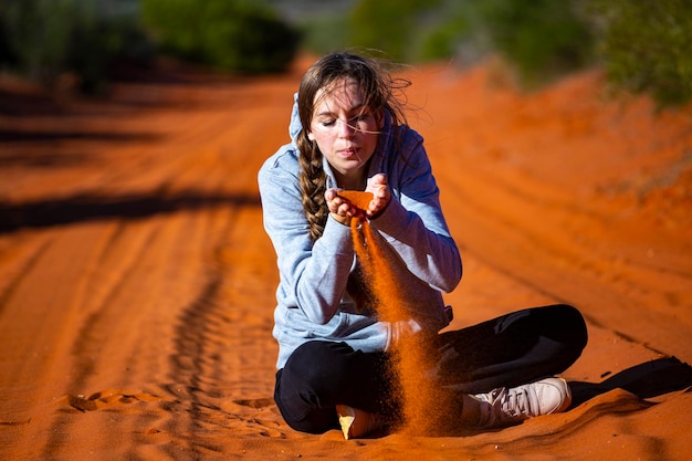 A beautiful long-haired girl plays with red sand on a sandy road in the desert