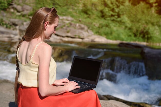 Beautiful long-haired girl in red skirt with laptop sitting on a rock on mountain river cascade. Freelance concept. Work in nature.