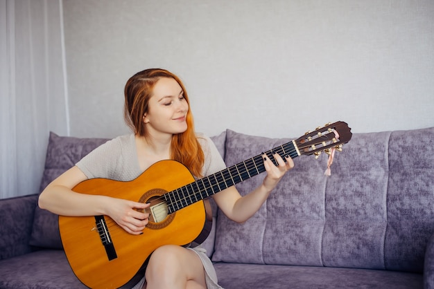 Beautiful long-haired young woman with positive mental health smiling, playing her favorite song on acoustic guitar, sitting on the couch at home. Musical hobby, free time.