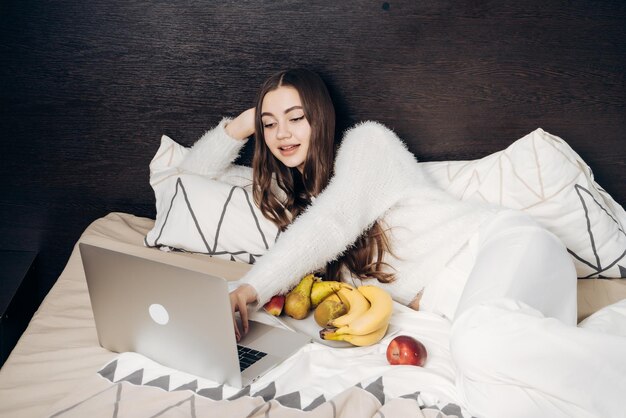 Beautiful longhaired girl in pajamas lies in bed late at night watching an interesting TV show and eating fruit