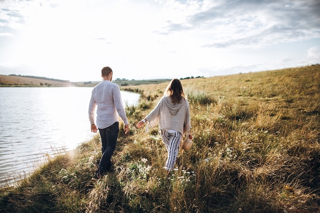 Beautiful loving couple walking on the shore, hugging, smiling on the sky background in the field. The guy and the girl hipster travel