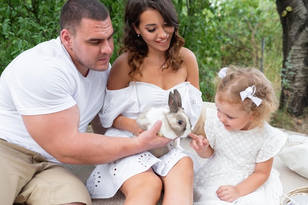 야외에서 즐거운 시간을 보내는 아름다운 사랑하는 가족 엄마 아빠와 어린 딸이 흰 토끼를 손에 들고 자연에 앉아 있습니다