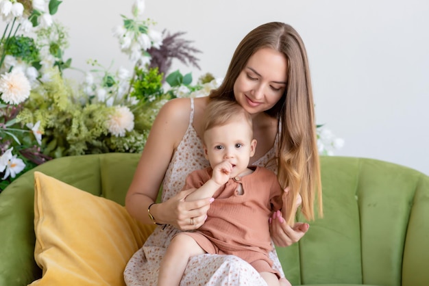 Beautiful loving mother in a dress sits on a green sofa surrounded by beautiful flowers She is holding her little son in a beige suit in her arms Happy loving family