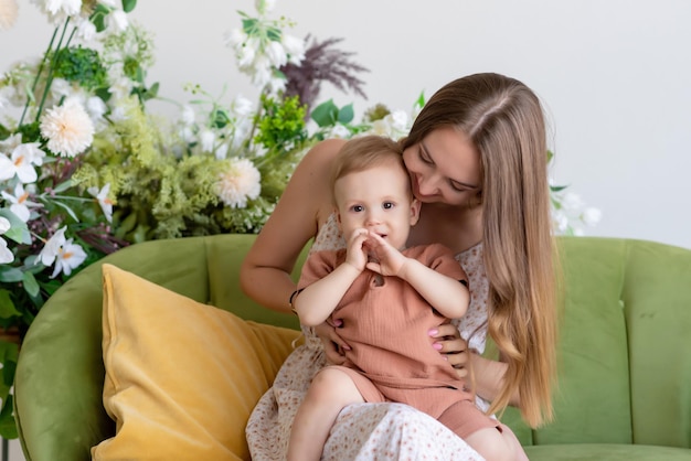 Beautiful loving mother in a dress sits on a green sofa surrounded by beautiful flowers She is holding her little son in a beige suit in her arms Happy loving family