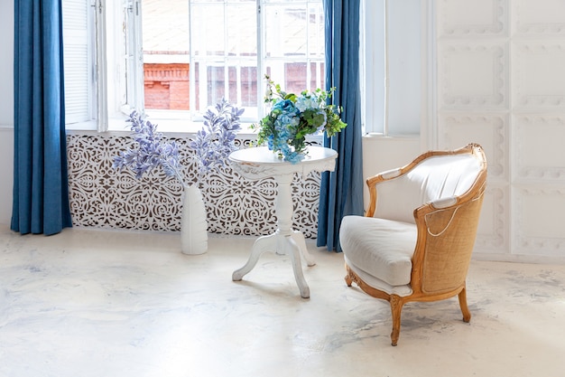 Beautiful luxury classic white bright clean interior bedroom in baroque style with large window, armchair and flower composition.