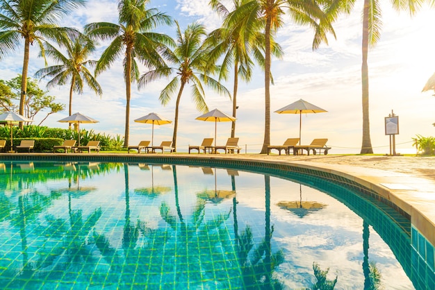 일몰 또는 일출 하늘에 코코넛 야자수가 있는 호텔 및 리조트의 야외 수영장 주변에 아름다운 고급 우산과 의자 - 휴일 및 휴가 개념