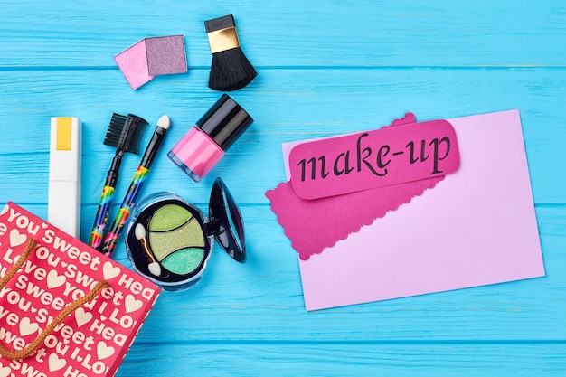 Beautiful make up cosmetics composition. Gift bag and cosmetics, blue wooden background. Pink card with text make-up, cosmetics items.