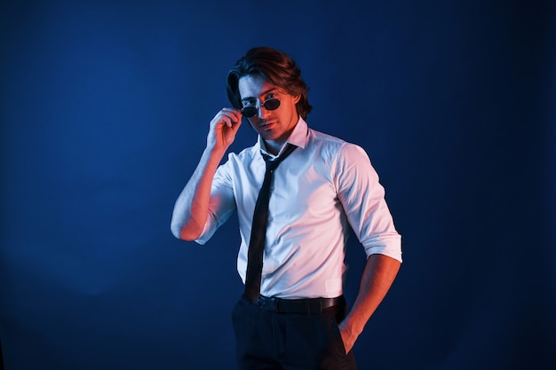 Beautiful man in formal wear and in glasses is in the studio with blue neon lighting.