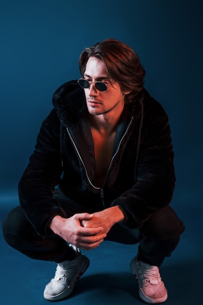 Beautiful man in glasses and black clothes sits in the studio with blue neon lighting.