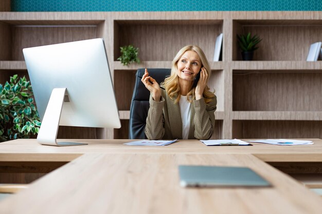 Beautiful mature adult businesswoman sitting at computer desk in the office and talking at phone