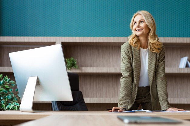 Beautiful mature adult businesswoman with elegant dress sitting at computer desk in the office