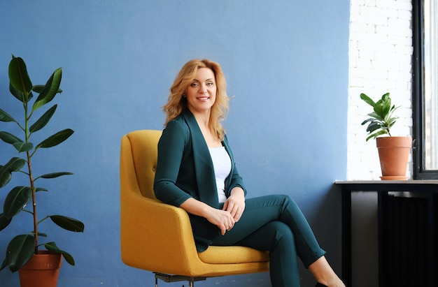 Beautiful mature blonde woman in a dark green trouser suit sits\
in a yellow armchair against a blue wall smiles and looks at the\
camera