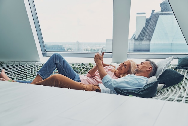 Beautiful mature couple examining smart phone and smiling while relaxing in big hammock at home toge...