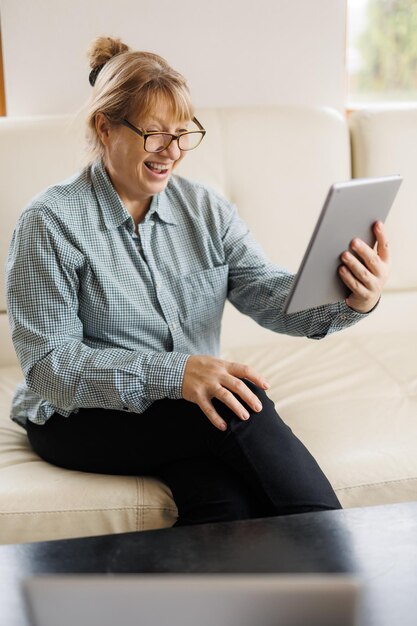 안경을 쓴 아름다운 성숙한 여성이 디지털 태블릿을 사용하고 집에 있는 소파에 앉아 웃고 있습니다.