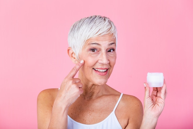 Vaso di detenzione bella donna matura di crema idratante e guardando la fotocamera. donna senior felice che tiene una bottiglia di lozione anti-invecchiamento isolata sopra fondo rosa. trattamento anti invecchiamento e bellezza.