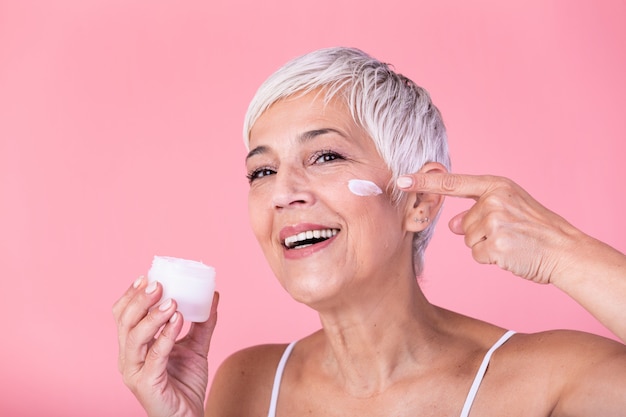 Beautiful mature woman holding jar of skin cream for face and body isolated on pink background. Happy senior woman applying anti-aging moisturizer and looking at camera. Beauty anti aging treatment.