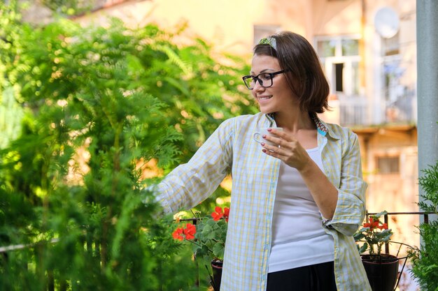 Beautiful mature woman in home clothes on open balcony with cup of coffee, smiling female looks away, in profile, copy space. Home balcony decorated with green plants and flowers