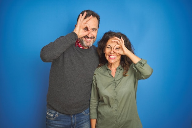 Beautiful middle age couple together standing over isolated blue background doing ok gesture with hand smiling eye looking through fingers with happy face