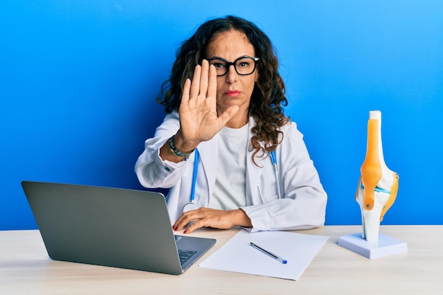 Beautiful middle age woman doctor at orthopedic clinic doing stop sing with palm of the hand warning expression with negative and serious gesture on the face