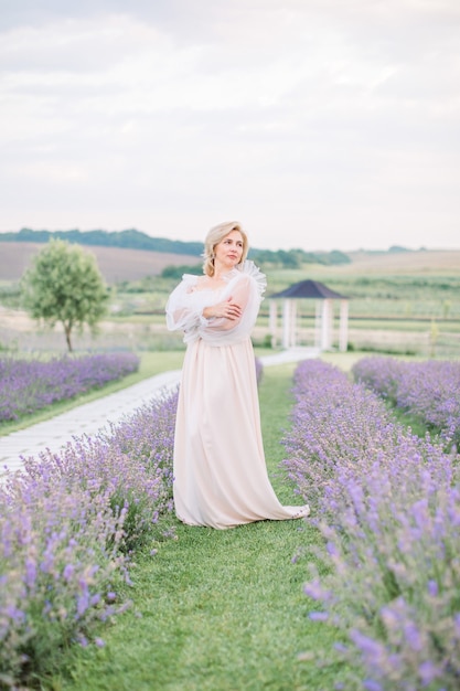 Beautiful middle aged blond Caucasian woman in a field of lavender. Woman in long elegant white dress walks on the lavender field.