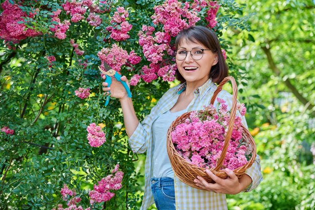 Beautiful middle aged smiling woman in garden caring for plants rose bush with flowers Happy mature female looking at camera in backyard Leisure hobby floriculture lifestyle nature 40s people