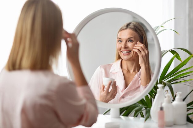 Фото Красивая женщина средних лет смотрит в зеркало и наносит увлажняющий крем для глаз