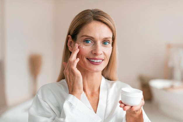 Photo beautiful middle aged woman in silky bathrobe using moisturizing eye cream enjoying face care routine