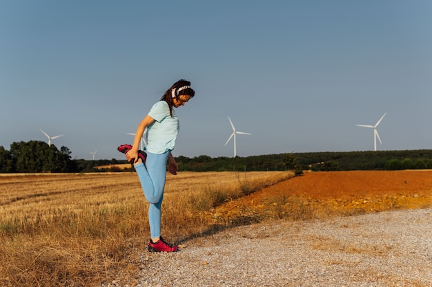 Beautiful middle-aged young woman stretching in the field outdoors, wearing music headphones with windmills on the horizon