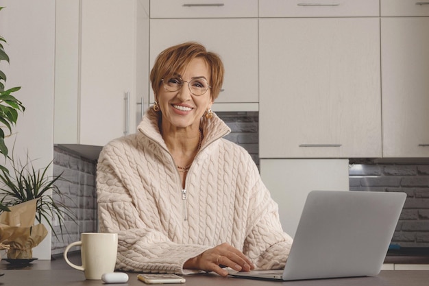 Beautiful middleaged baby boomer woman working in laptop and happy smiling background of light gray ...