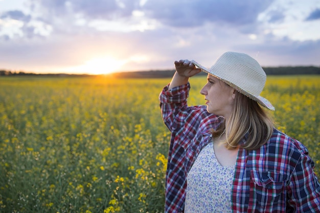 A beautiful middleaged farmer woman in a straw hat and a plaid shirt stands in a field of flowering rapeseed in the sunlight