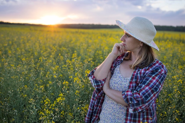 A beautiful middleaged farmer woman in a straw hat and a plaid shirt stands in a field of flowering rapeseed in the sunlight