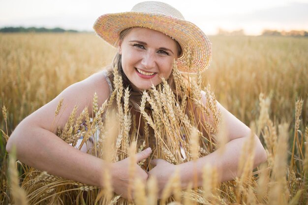 A beautiful middleaged farmer woman in a straw hat and a plaid shirt stands in a field of golden ripening wheat during the daytime in the sunlight