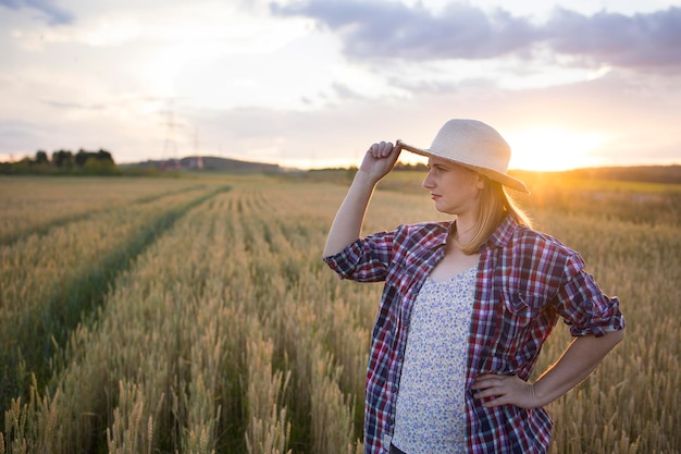 A beautiful middleaged farmer woman in a straw hat and a plaid shirt stands in a field of golden ripening wheat during the daytime in the sunlight