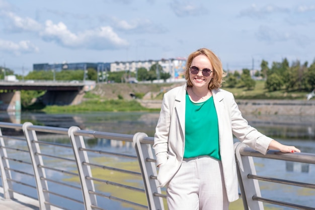 A beautiful middleaged woman in a suit jacket is standing on the bank of the river with her hand in her pocket