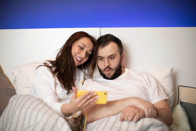 Beautiful millennial couple using mobile phone lying on bed together