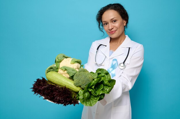 Bella nutrizionista donna di razza mista in abito medico e nastro blu di sensibilizzazione sul diabete posa con piatto di cibo crudo sano su uno sfondo blu con copia spazio per la pubblicità medica