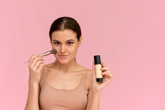 a beautiful model applies foundation with a brush to clean skin on pink background